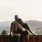 A woman leans on a man's shoulder while he holds her; they are standing on a deck overlooking the Blue Ridge Mountains.