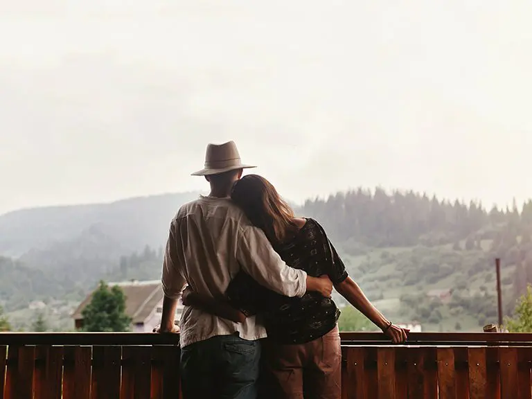 A woman leans on a man's shoulder while he holds her; they are standing on a deck overlooking the Blue Ridge Mountains.