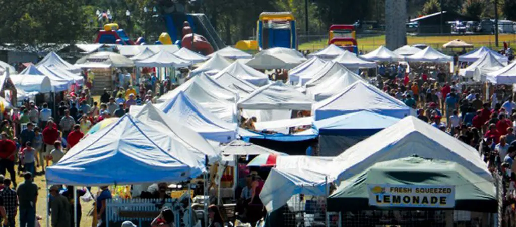 A bustling view of the Ellijay Georgia Apple Festival with rows of vendor tents, large crowds browsing food and crafts, and family attractions in the background.