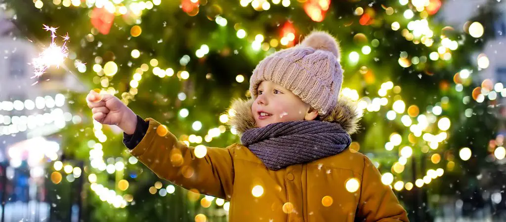 Child holding a sparkler in front of a lit Christmas tree on a snowy day
