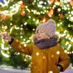 Child holding a sparkler in front of a lit Christmas tree on a snowy day