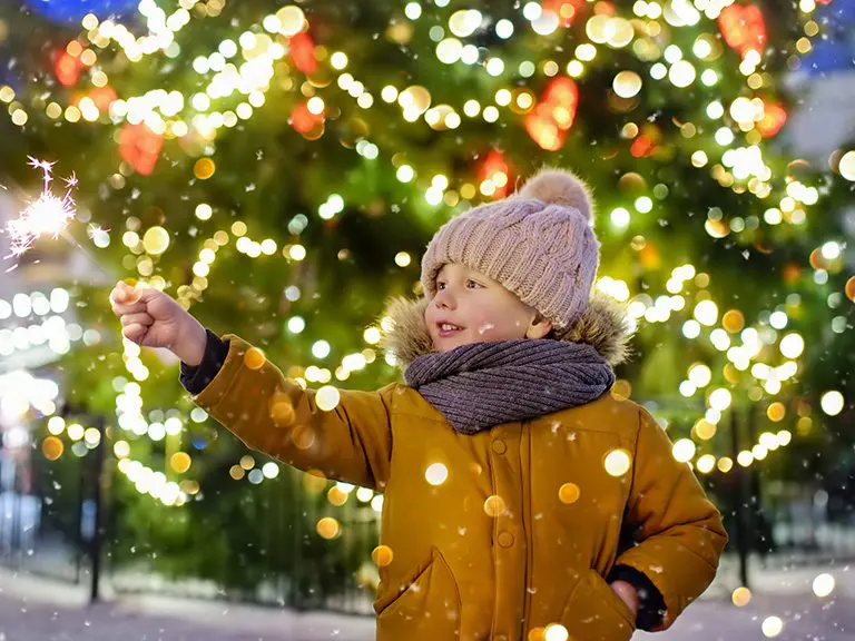 Child holding a sparkler in front of a lit Christmas tree on a snowy day