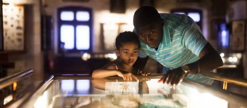 A dad and daughter lean over a backlit display case in a museum, pointing to artifacts. 