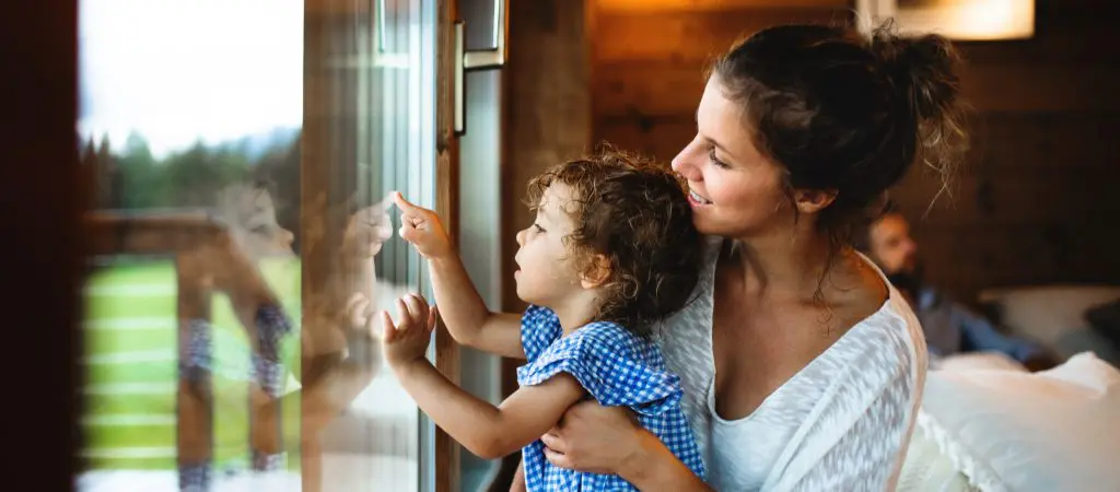 A mom holds her little girl up to see nature outside the window of a log cabin, with the dad lounging on the couch in the background.