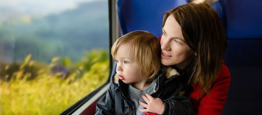 Child sitting in their mom's lap looking out a train window at fields and mountain scenery with awe.