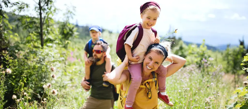 A little girl sitting on Mom's shoulders while smiling and hiking through a sunny wildflower field among a forested area, with her younger brother on Dad's shoulders and mountains in the background.