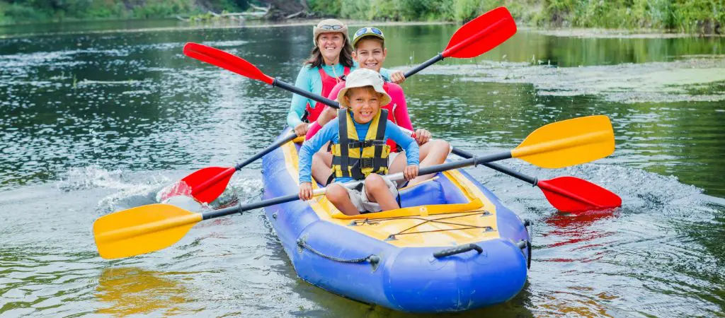 A mom, teenager, and small boy paddle  in an inflatable canoe along a tree-shaded river, each smiling with happiness and excitement.