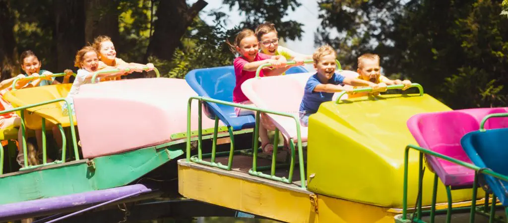 Many children smile big while riding a roller coaster over a hill, with trees in the background.