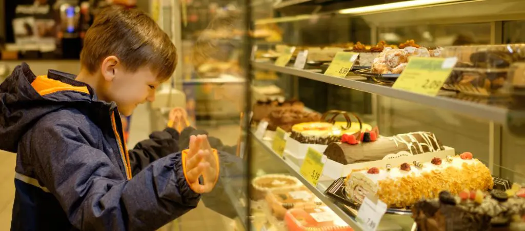 A little boy in a jacket leans with both hands and a big grin to peer into the glassed sweets cabinet.