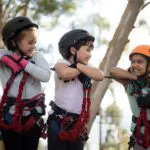 Three children in zipline gear leaning on a rope bridge in the treetops, each grinning ear to ear.