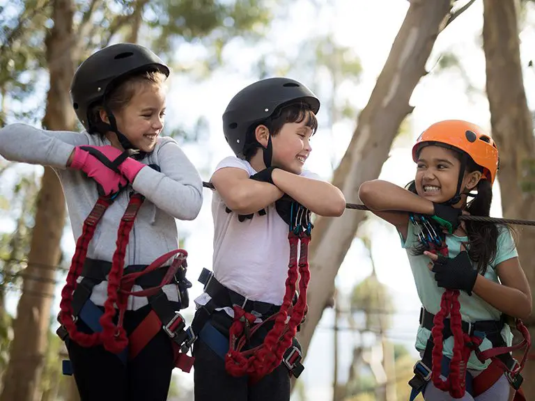 Three children in zipline gear leaning on a rope bridge in the treetops, each grinning ear to ear.