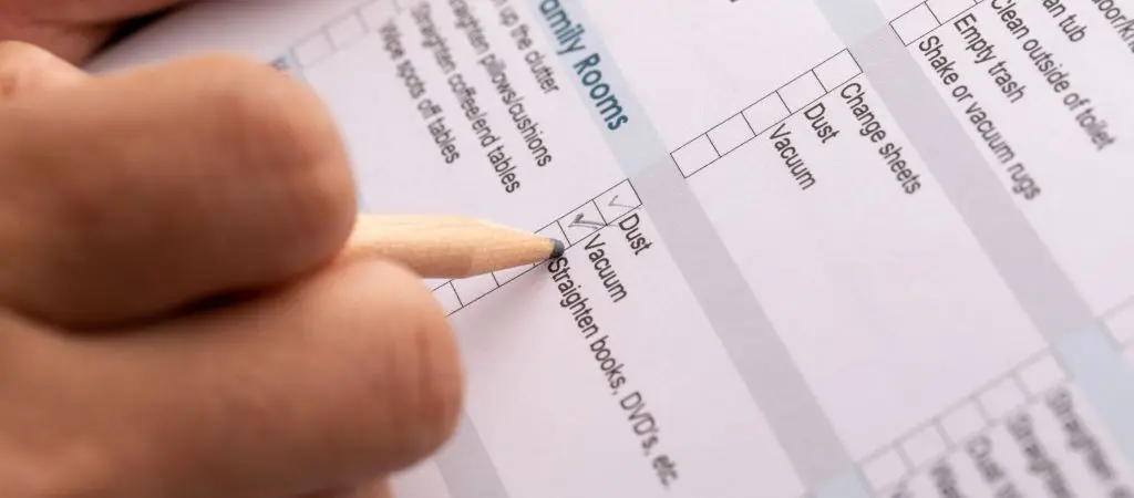 Hand marking off squares on a cleaning checklist, like dust, vacuum, and straighten DVDs, with a pencil
