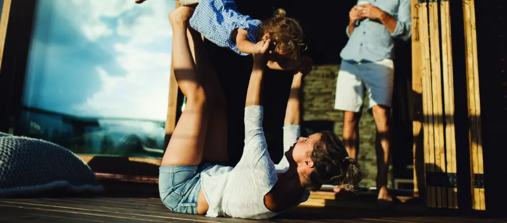 Mom holding up child in the air while laying on a cabin deck, with the father onlooking and mountain views in the reflection of a large picture window