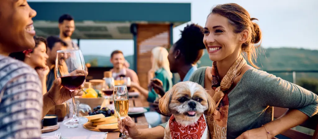 A woman with a small dog in her lap smiles with a group of friends while sitting an outdoor table, with mountains in the background.