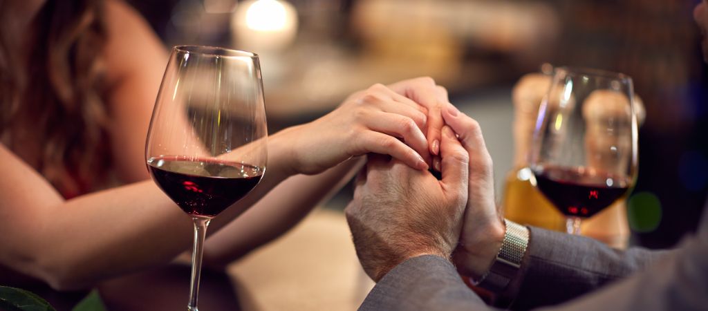 A couple hold hands across a table with 2 wine glasses.