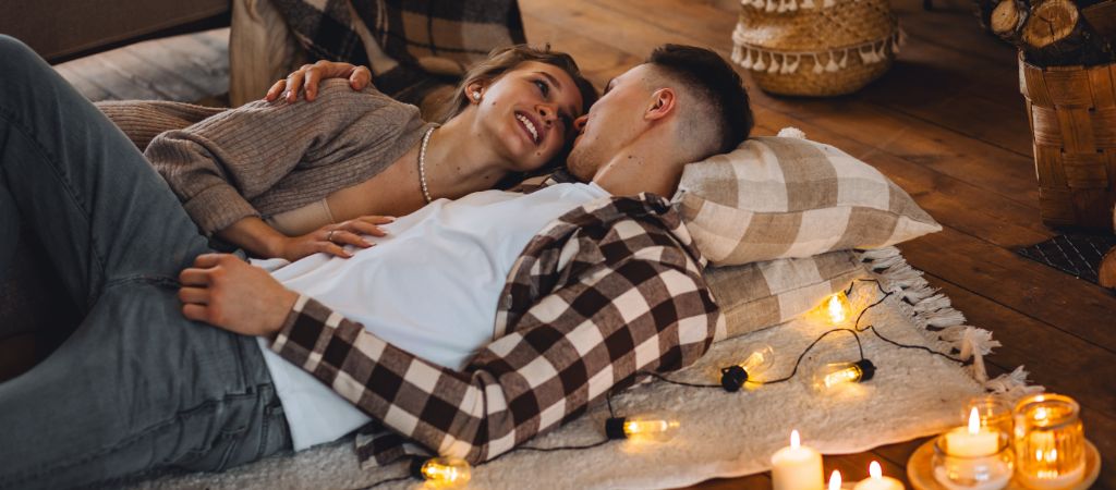 A young couple lays on pillows and a blanket spread on the wooden floor of a log cabin while cuddling and smiling at each other, with candles and string lights illuminating the space.