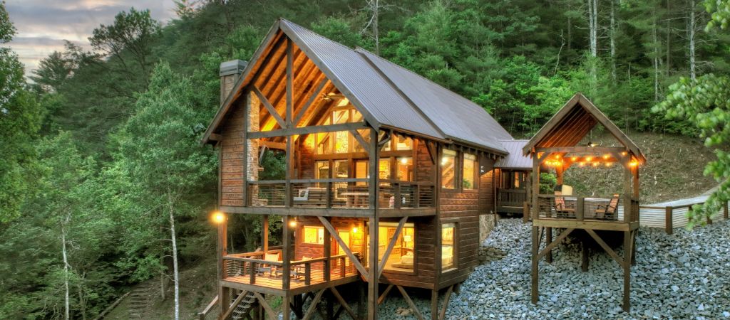 A large Blue Ridge Mountain cabin surrounded by trees lights up in the evening, with a covered hot tub on the side.