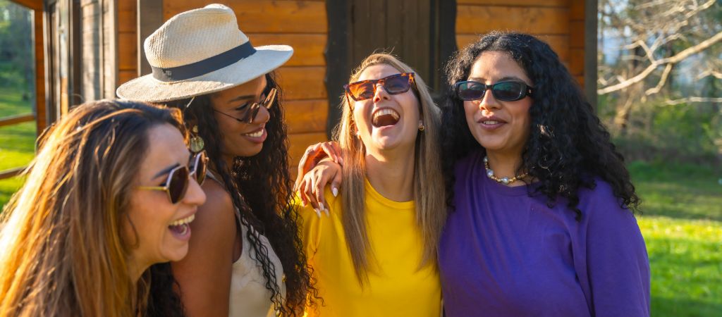 Four women smile and laugh together on a sunny day in front of a log cabin.