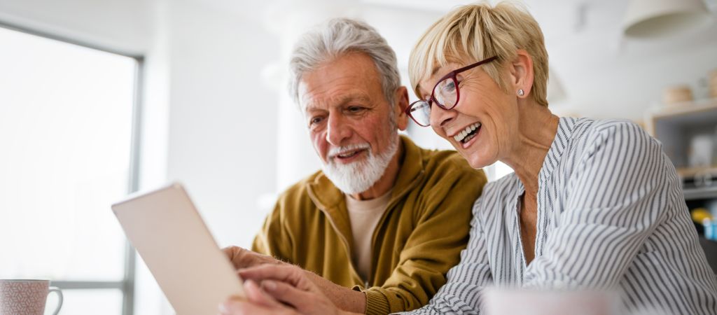 A senior couple smile while pointing to a tablet screen inside their home.