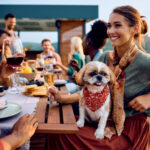 A woman with a small dog in her lap smiles with a group of friends while sitting an outdoor table, with mountains in the background.