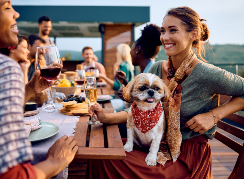 A woman with a small dog in her lap smiles with a group of friends while sitting an outdoor table, with mountains in the background.
